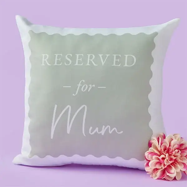 A soft square cushion with a scalloped border design and the words Reserved for Mum on the front, displayed beside a pink flower on a pastel purple background.